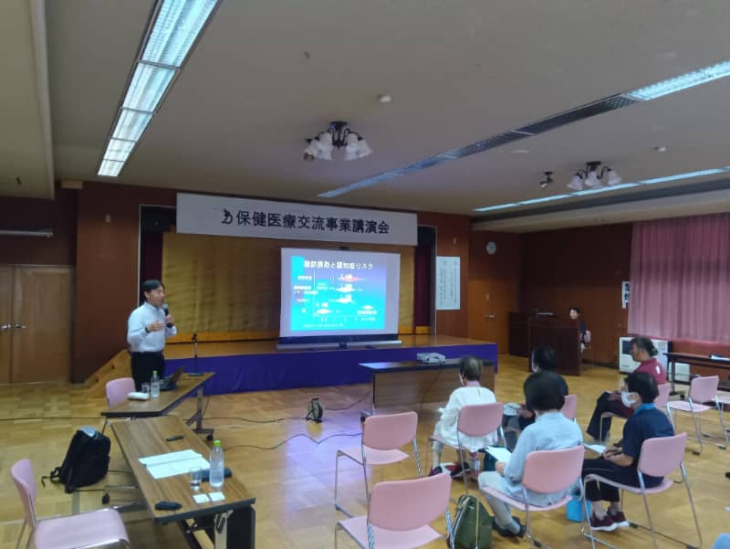 講演会で大平哲也教授がスライドを使いながら「笑いで心も体も健康に」について話している様子。参加者は座って聴講中。