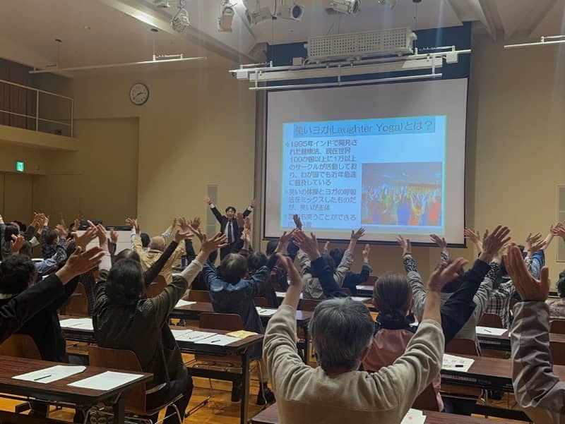 笑いヨガの講演会で参加者が手を挙げている様子、スクリーンには関連情報が表示されている。