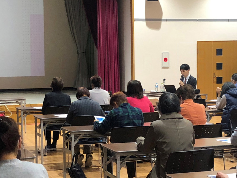 講演会で話をする男性と、聴衆が座っている様子。参加者はメモを取ったり、資料を見たりしている。