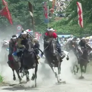 福島の伝統的な馬追い祭りで、騎手たちが馬に乗って競い合う様子。観客が見守る中、砂煙が舞い上がる。
