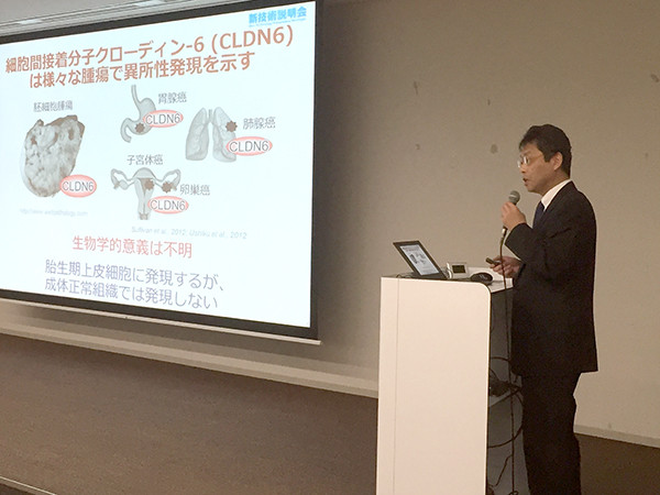 スーツ姿の男性がマイクを持ちスライドを用いて講演している