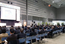 多くの人が前方のスクリーンと演台に注目して座っている様子