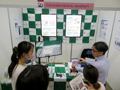 福島医科大学の展示ブースで、医療技術について説明を受けている人々。