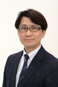 福島県立医科大学の腫瘍内科学講座主任教授、佐治重衡氏のポートレート。白衣を着用し、背景には書類や小物が並ぶ。