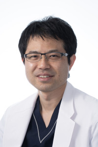 福島県立医科大学の腫瘍内科学講座主任教授、佐治重衡氏のポートレート。白衣を着用し、背景には書類や小物が並ぶ。