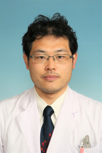 福島県立医科大学の腫瘍内科学講座主任教授、佐治重衡氏のポートレート。白衣を着用し、背景には書類や小物が並ぶ。