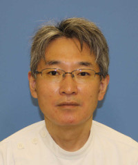 福島県立医科大学の腫瘍内科学講座主任教授、佐治重衡氏のポートレート。白衣を着用し、背景には書類や小物が並ぶ。