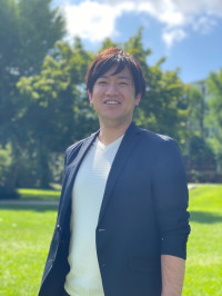 福島県立医科大学の腫瘍内科学講座主任教授、佐治重衡氏のポートレート。白衣を着用し、背景には書類や小物が並ぶ。
