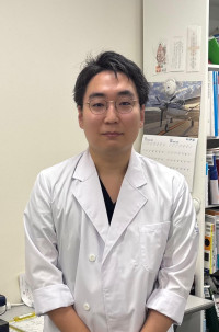 福島県立医科大学の腫瘍内科学講座主任教授、佐治重衡氏のポートレート。白衣を着用し、背景には書類や小物が並ぶ。