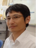 福島県立医科大学の腫瘍内科学講座主任教授、佐治重衡氏のポートレート。白衣を着用し、背景には書類や小物が並ぶ。