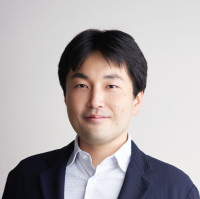 福島県立医科大学の腫瘍内科学講座主任教授、佐治重衡氏のポートレート。白衣を着用し、背景には書類や小物が並ぶ。