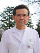 福島県立医科大学の中村常幸氏が白衣を着て微笑んでいる写真。背景には緑の木々が見える。
