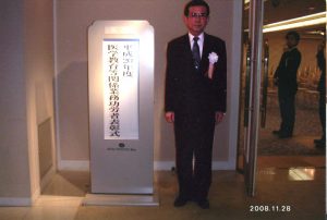 解剖実習に係る献体業務で表彰された男性が、受賞式の看板の前に立っている写真。