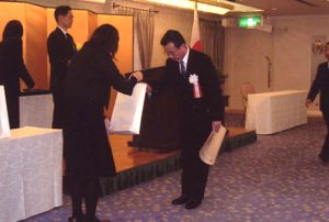 表彰式で賞を受け取る男性と、賞状を手渡す女性が写っているシーン。背景には日本の国旗が見える。