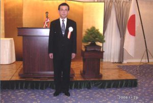 福島県立医科大学の表彰式で、スーツを着た男性が壇上に立ち、背景には日本の国旗と盆栽がある。