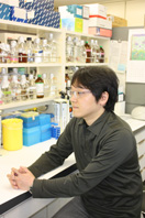 研究室で考え込む男性の横顔と、背後に並ぶ試薬瓶や実験器具が映る。