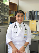 川崎幸彦准教授が医療現場で白衣を着て座っている写真。背景には書類や医療器具が見える。