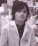 福島県立医科大学の微生物学講座の講師、生田和史氏が白衣を着て研究室でポーズをとっている写真。