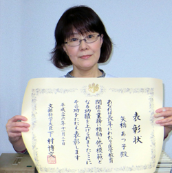 医療技師としての功績を表彰された女性が、賞状を手に微笑んでいる様子。