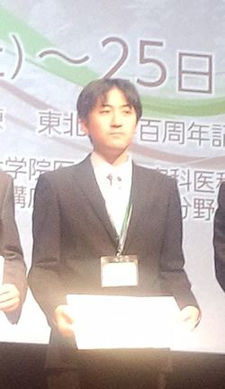 学会で表彰状を持つ男性がスーツ姿で立っている様子。背景にはイベントのタイトルが表示されている。