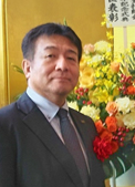 厚生労働大臣表彰を受けた山寺幸雄技師長が花々の前で微笑む姿。