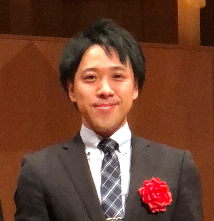 医療技師の男性がスーツを着て微笑んでいる写真。胸元には赤い花のコサージュが付いている。