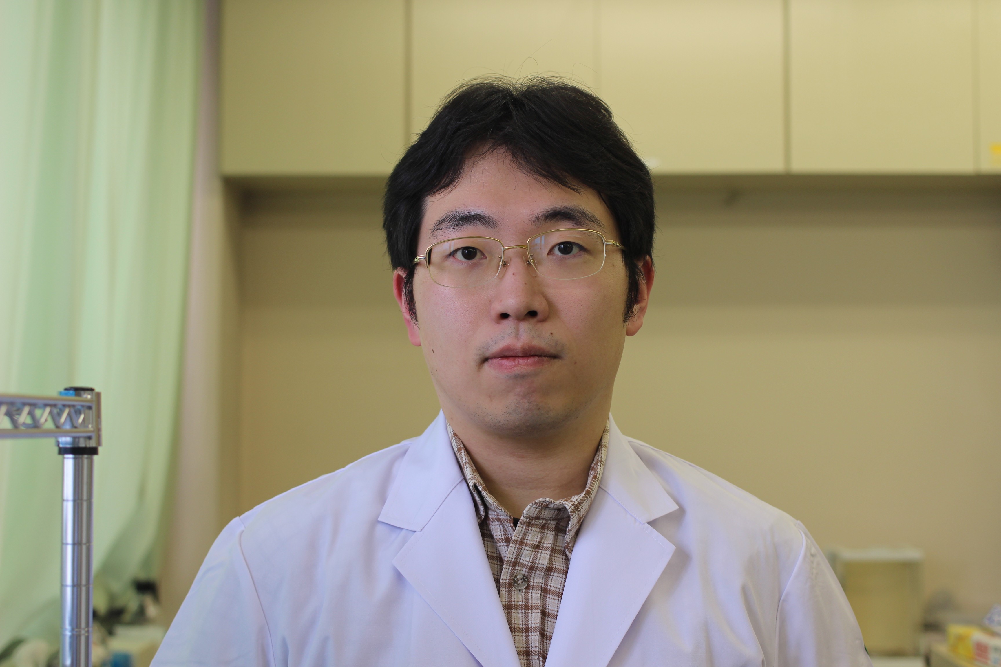 医学部の白衣を着た男性が、研究室と思われる場所でカメラを見つめている。背景には実験器具が見える。