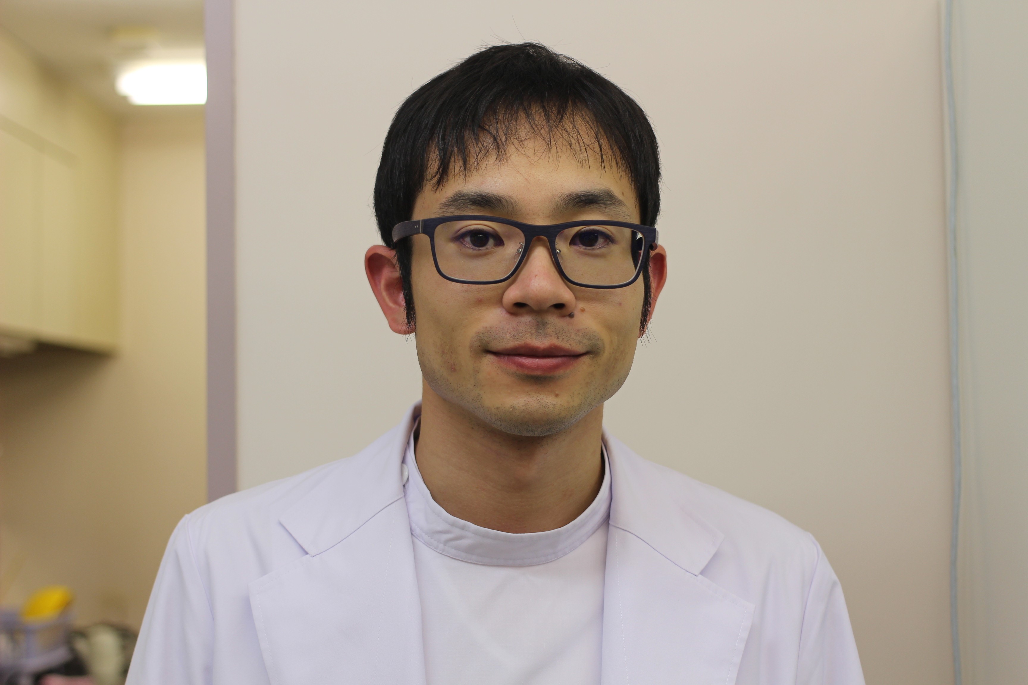 医療用白衣を着た男性がカメラに向かって微笑んでいるポートレート。背景には医療施設の一部が見える。