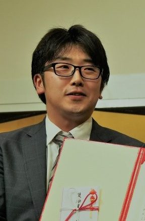 重症HUSモデルマウスの研究に関する表彰を受けた男性が、賞状を手に微笑んでいる様子。