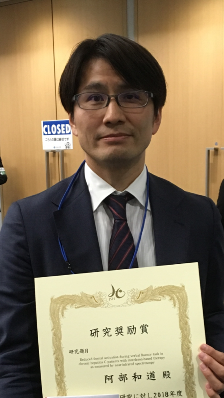 研究奨励賞を持つ阿部和道講師が微笑む姿。背景には「CLOSED」のサインが見える。