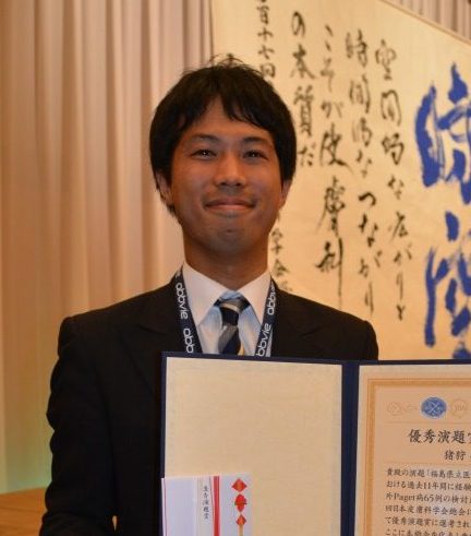 福島県立医科大学で表彰を受けた男性が、賞状を手に微笑んでいる写真。背景には書道作品が見える。