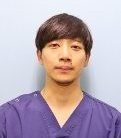 医療用の服を着た男性がカメラに向かって微笑んでいるポートレート。背景は淡い青色。