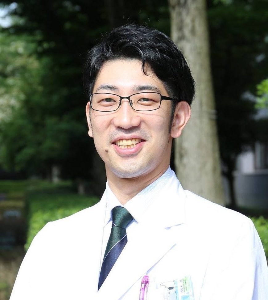 医療白衣を着た男性が笑顔で立っている写真。背景には緑の木々が見える。