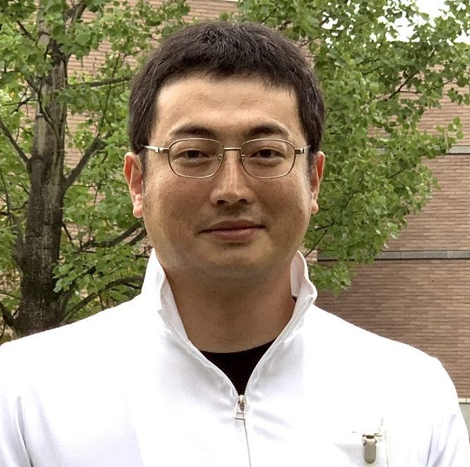 福島県立医科大学の小島学博士が白衣を着て微笑んでいるポートレート。背景には緑の木々が見える。