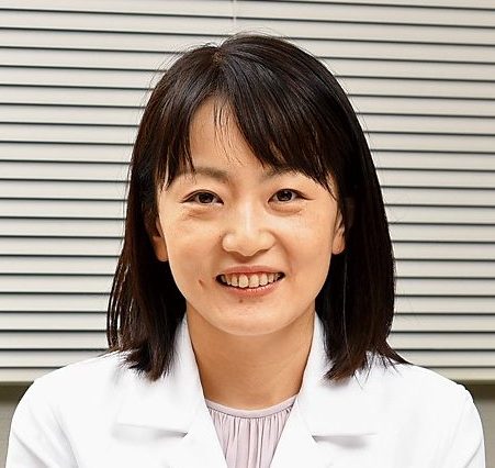 医療用白衣を着た女性が微笑んでいるポートレート。背景にはブラインドが見える。