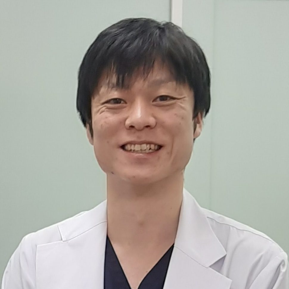 医療用白衣を着た男性が微笑んでいるポートレート。背景は淡い色合いの壁。