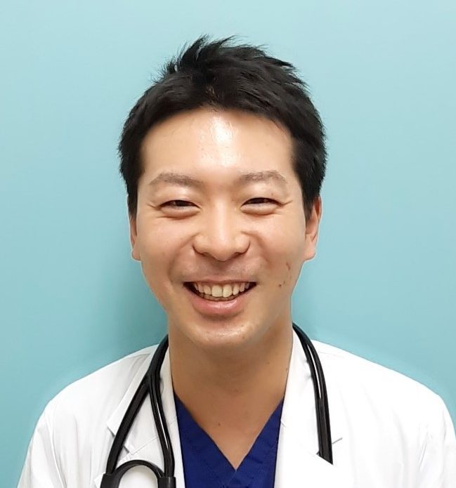 医療用白衣を着た男性が笑顔でカメラを見つめている写真。背景は淡い青色。