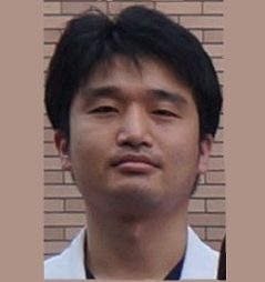 医療白衣を着た男性が、背景のレンガ壁の前で真剣な表情を浮かべている。