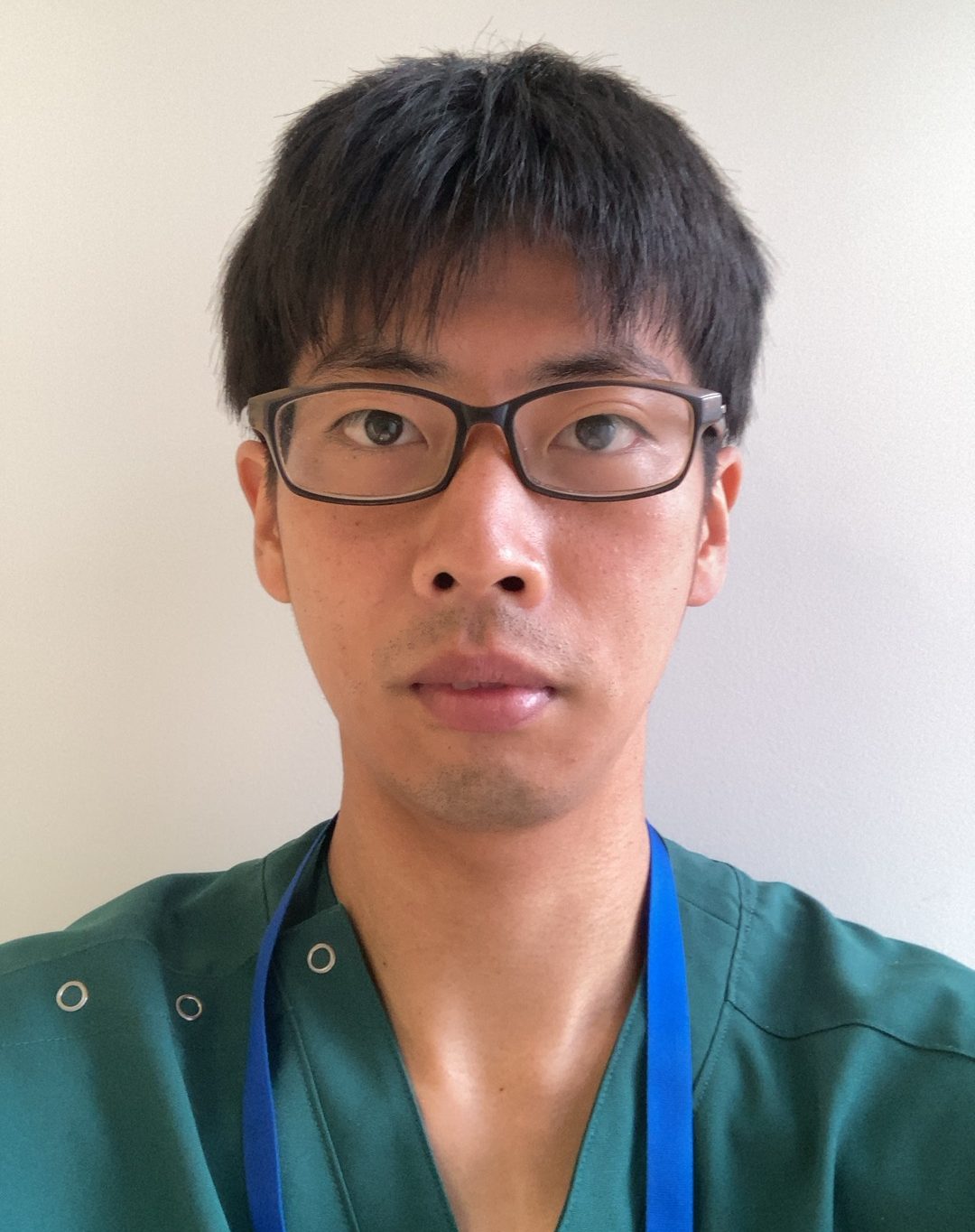 医療用の服を着た男性がカメラを見つめている自撮り写真。背景は無地の壁。