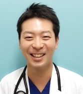 医療用白衣を着た男性が笑顔でカメラを見つめているポートレート。背景は淡い青色。