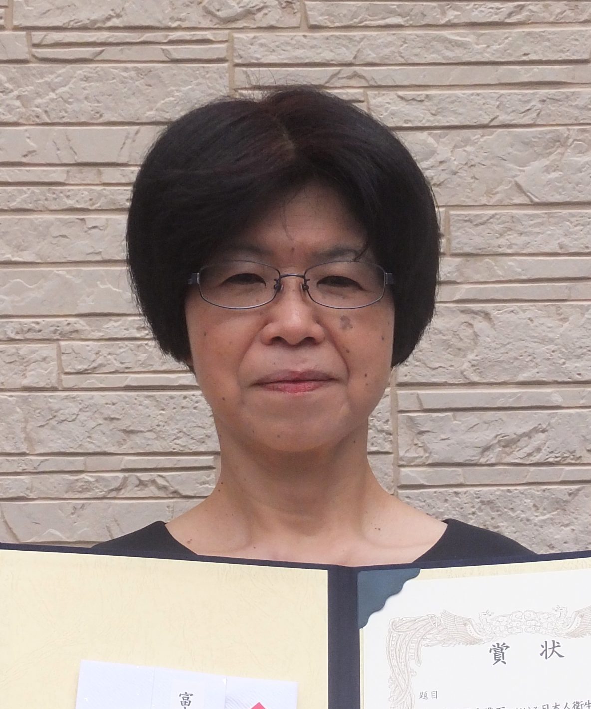 日本の衛生学者が賞状を持って微笑んでいる写真。背景は淡い色の壁。