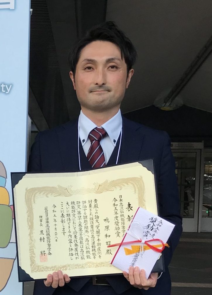 医療技師の男性が表彰状を持ち、笑顔でポーズをとっている写真。