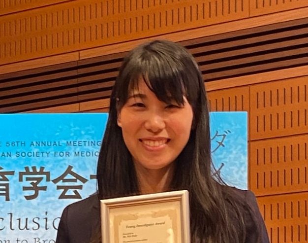 学生が表彰状を持って微笑む姿、背景には医療教育に関するイベントの看板が見える。