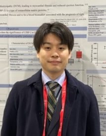 福島県立医科大学の西浦司人助手が研究発表のポスターの前で微笑んでいる写真。