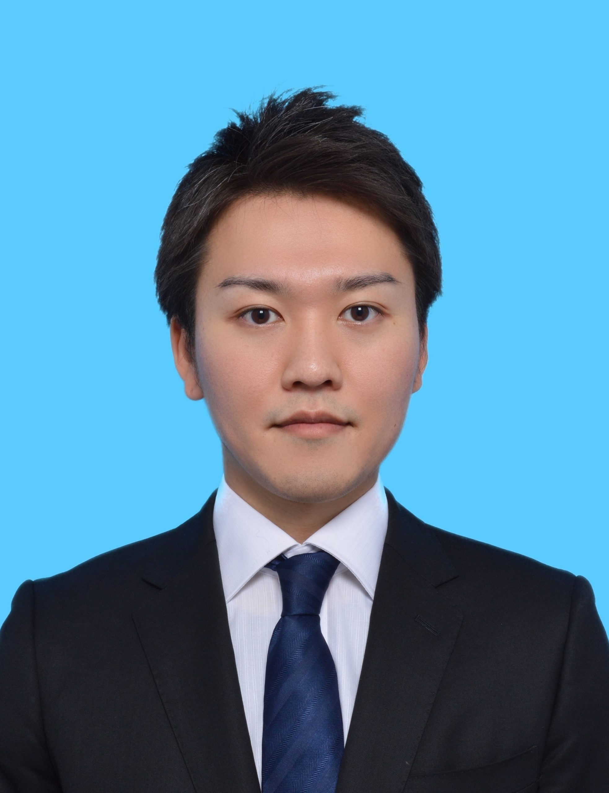 近藤悠斗助教のプロフィール写真。青い背景の前でスーツを着た男性のポートレート。