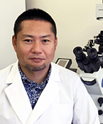 井上直和准教授が顕微鏡の前で微笑む姿。医学研究に従事する専門家の様子。