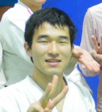 笑顔でピースサインをする男性が写っている写真。背景には他の人々がいる。