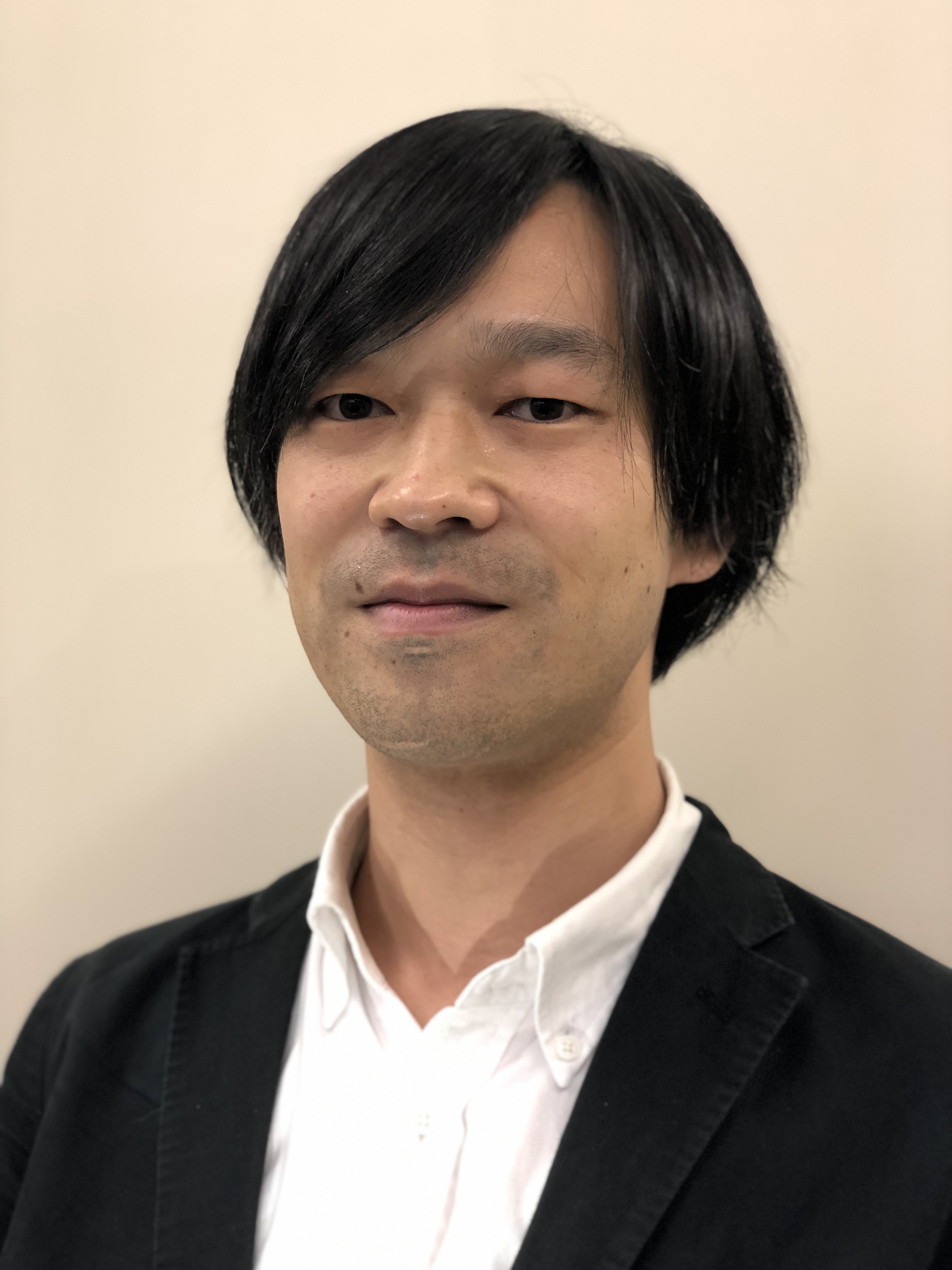 黒いジャケットを着た男性が微笑んでいるポートレート。背景は淡い色合いの壁。