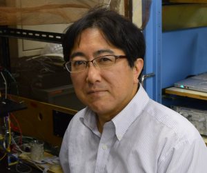 挾間章博教授が研究室で微笑む姿。背景には研究機器が配置されている。