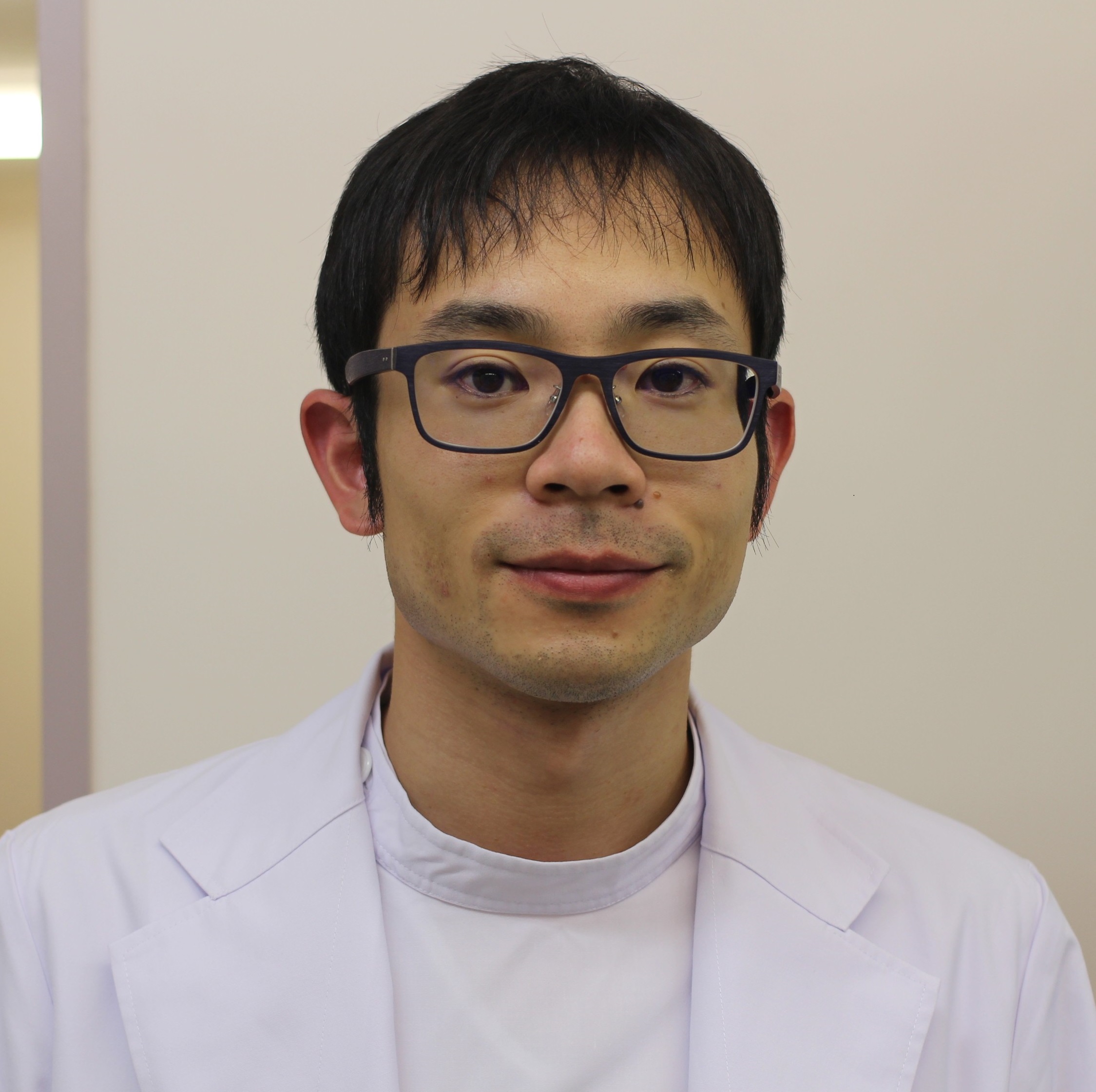 医療用白衣を着た秦淳也講師がカメラに向かって微笑んでいるポートレート。